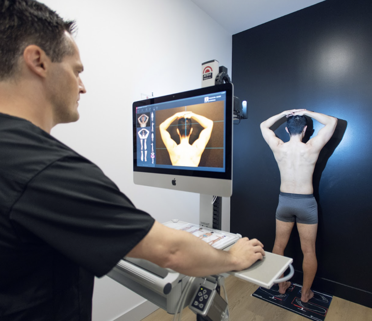 A clinician operates a full-body photography system while a patient poses for imaging.