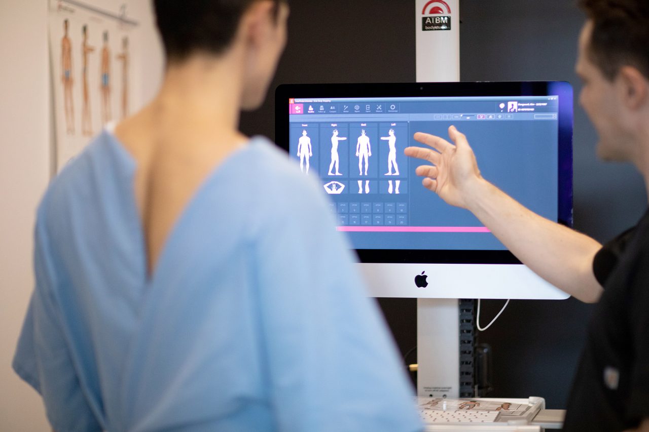 A clinician points to body photographs on a mole-mapping monitor.