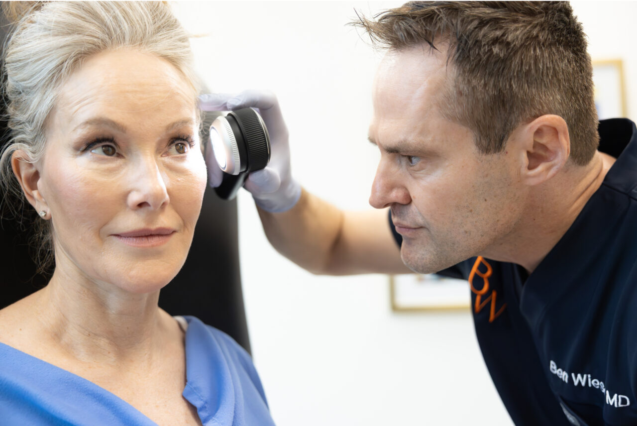 A clinician examines a patient's forehead with a dermatoscope.