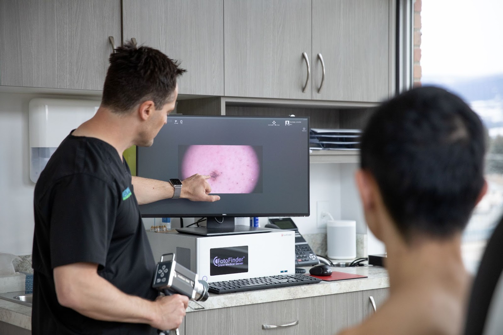 A clinician and patient review a magnified skin image on a monitor.