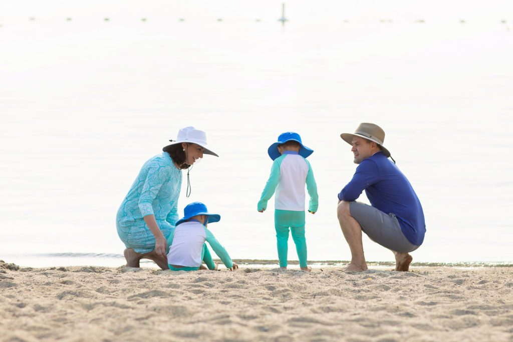 Mother, Father and two young children at the beach all wearing UV protective clothing that if worn regularly, will protect them from the damages caused by continued sun exposure.