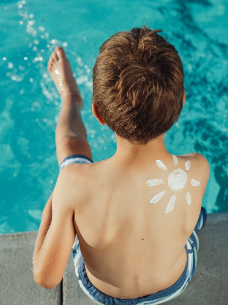 Young boy sitting on the edge of a pool with a sun drawn on his back in sunscreen.