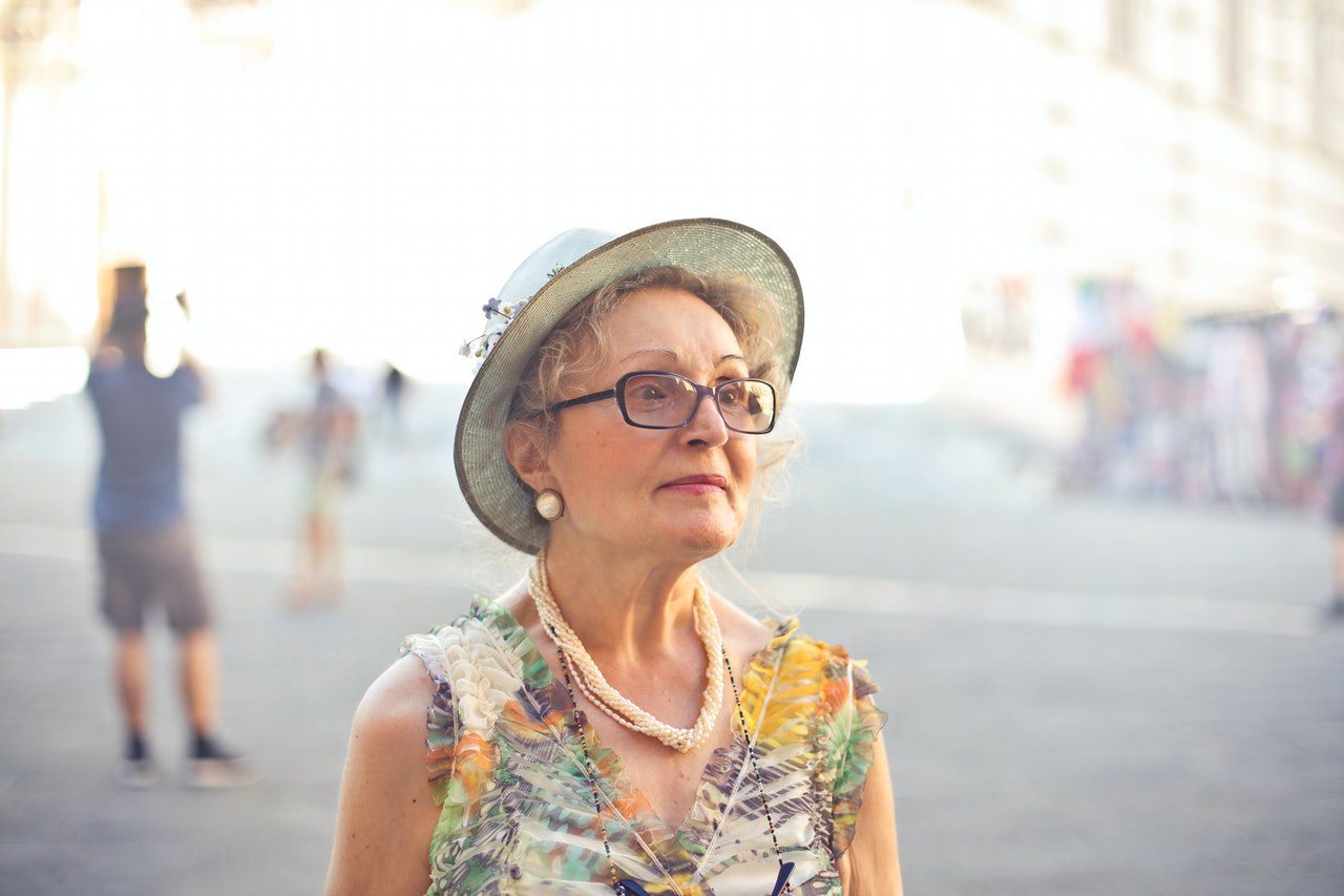 a senior woman standing outside in a market, wearing black-rimmed glasses, a bright blouse and brimmed hat