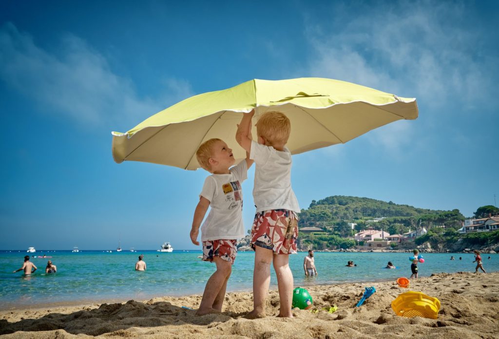 Two preschool aged boys with blonde hair at the beach under a yellow beach umbrella.