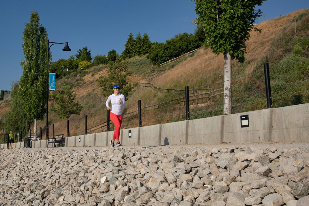 Woman in athletic clothing jogging on a path by the lake in West Kelowna, relating to getting more sun to produce vitamin D to prevent melanoma.